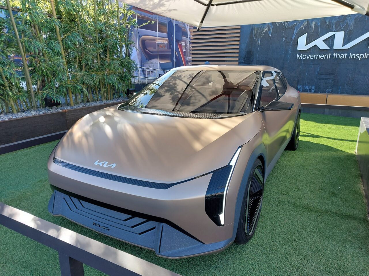 Projekty samochodów CES 2024. Koncept elektrycznego samochodu marki KIA wystawiony na zewnątrz na sztucznej trawie, z bambusami w tle i napisem "Movement that inspires".