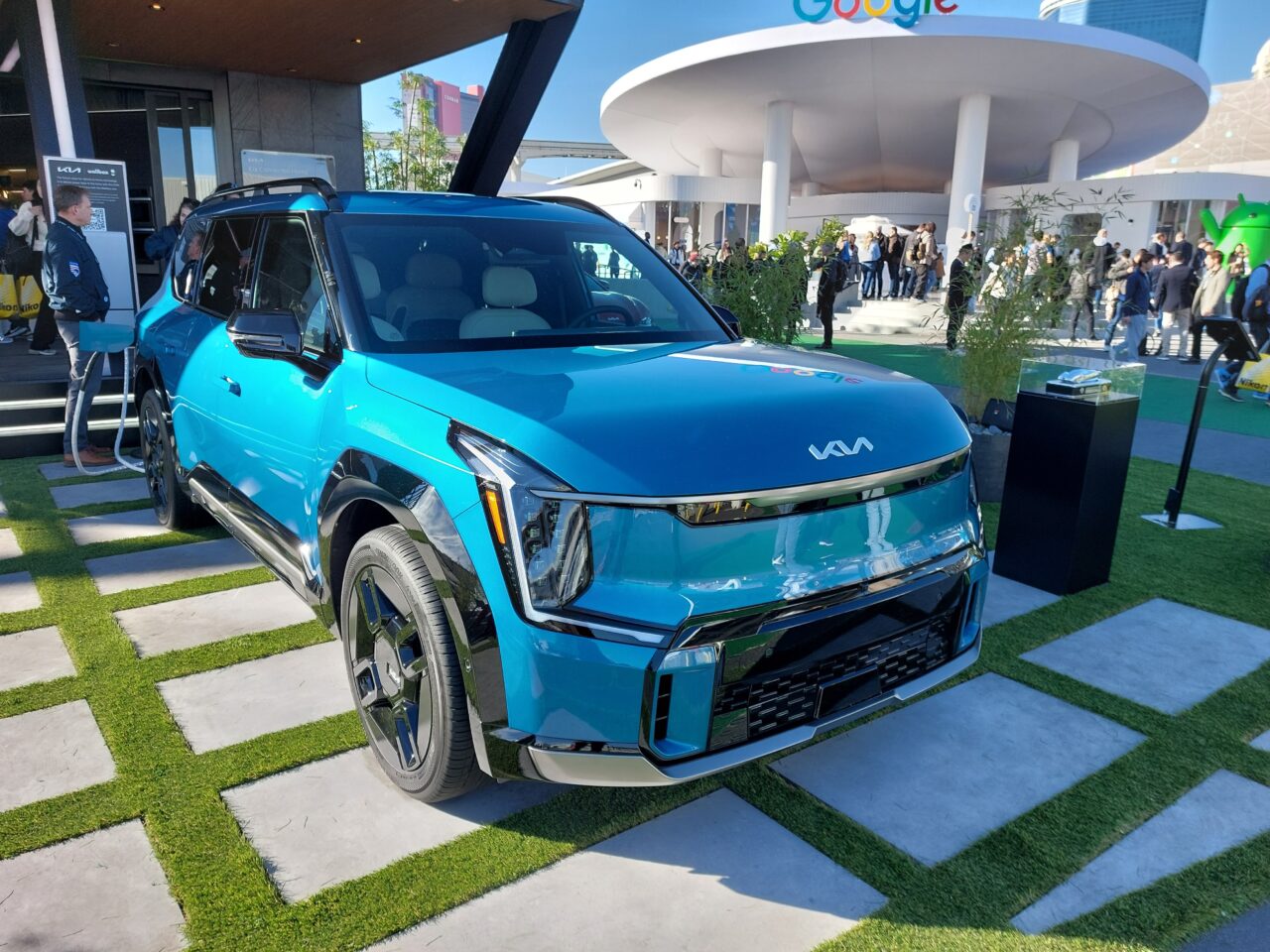 Niebieski samochód elektryczny marki Kia zaparkowany na sztucznej trawie z logo Google w tle i grupą ludzi.
