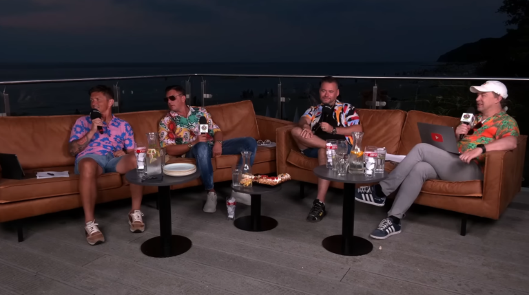 Prowadzący Kanał Sportowy. Czterech mężczyzn siedzących na brązowej kanapie na zewnątrz z mikrofonami, napojami i widokiem na morze w tle.