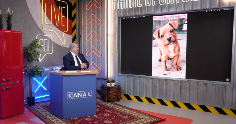 Prezenter telewizyjny siedzący za pulpitem w studio, śmiejący się na widok wyświetlanego na ekranie obrazu szczeniaka z napisem "monkey and a rabbit".