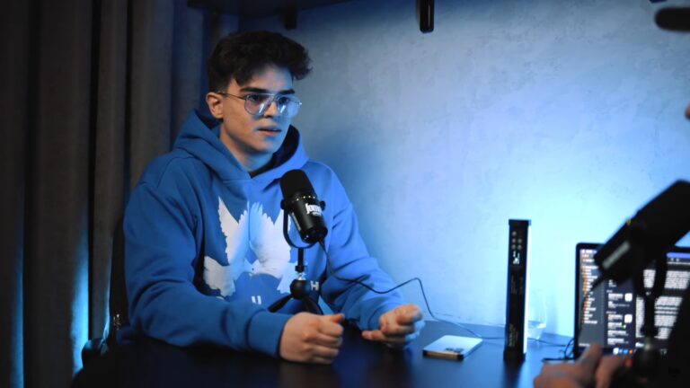 Młody mężczyzna w niebieskiej bluzie z kapturem siedzi przed mikrofonem w podświetlonym na niebiesko pokoju, wydaje się być w trakcie nagrywania podcastu lub rozmowy.