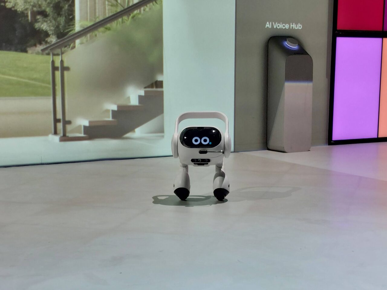 Mały biały robot LG AI Agent z dwoma czarnymi oczami na cyfrowym wyświetlaczu stoi na jasnej posadzce przed kolorowymi ekranami i urządzeniem oznaczonym napisem "AI Voice Hub".
