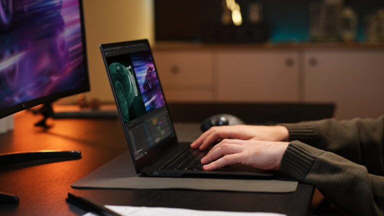 Osoba używająca laptopa z grafiką 3D samochodu na ekranie, znajdująca się przy biurku z dodatkowym monitorem w tle wyświetlającym dynamiczny obraz samochodu.