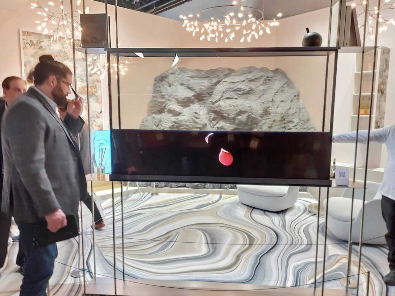 Przezroczysty telewizor LG Signature OLED T, w tle sztuczne skały, widoczni ludzie i elementy dekoracyjne wnętrza na stoisku.
