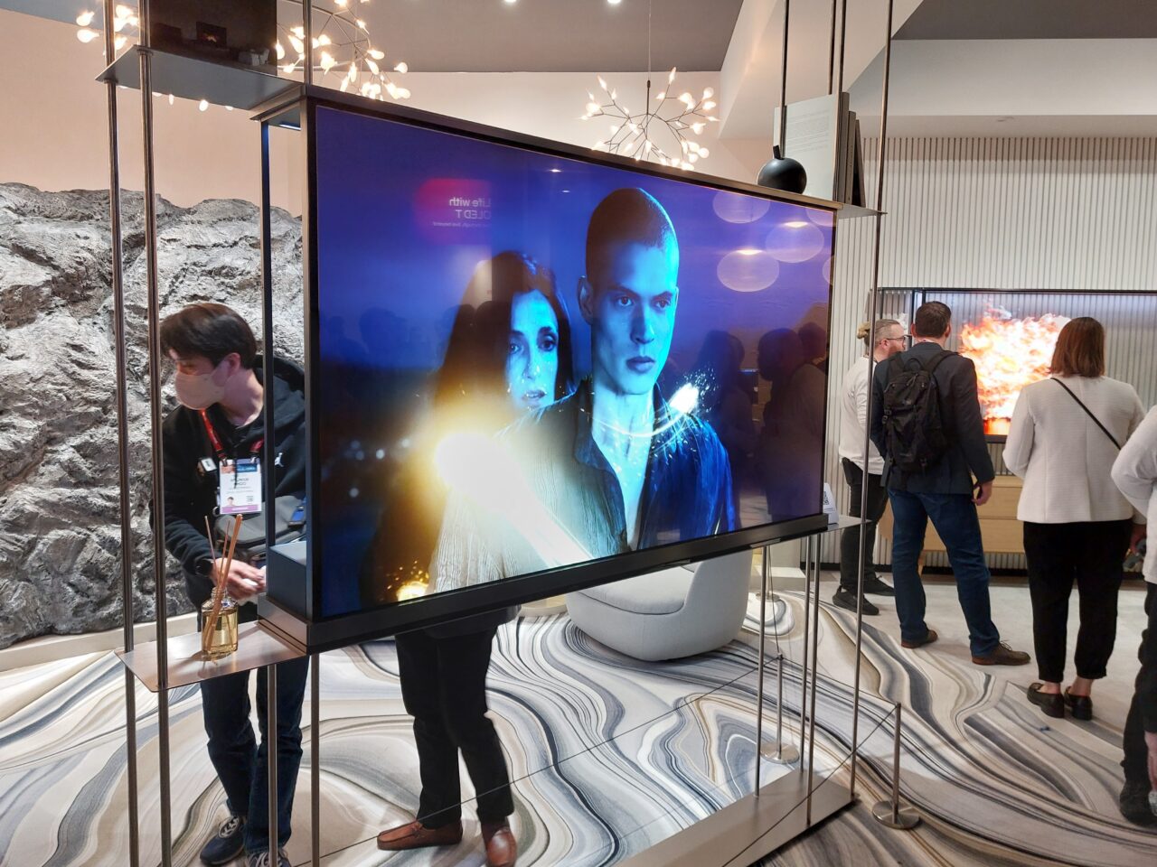Telewizor LG Signature OLED T na stojaku prezentujący grafikę z dwoma postaciami, w tle widoczni ludzie zwiedzający pokaz technologiczny.