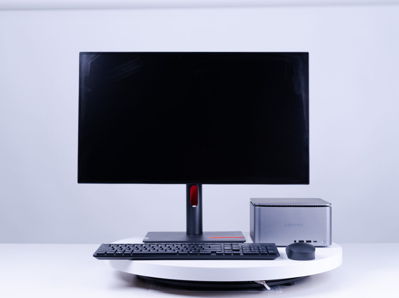 Monitor komputerowy, klawiatura, mini PC marki Lenovo i mysz na białym biurku przed jasnym tłem.