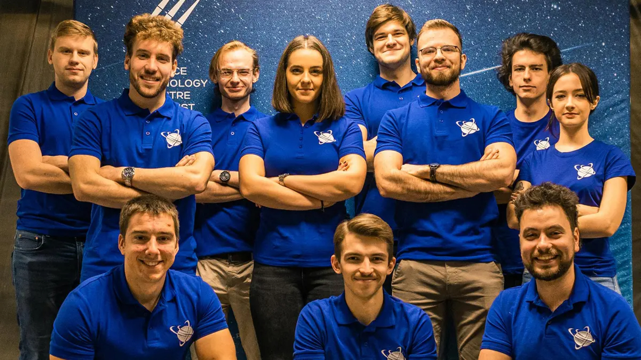 koło naukowe AGH Lunar Technologies stojące za projektem Lunaris w jednolitych niebieskich koszulkach z logotypem, ustawionych w dwóch rzędach na tle o tematyce kosmicznej, z uśmiechniętym mężczyzną klęczącym z przodu.
