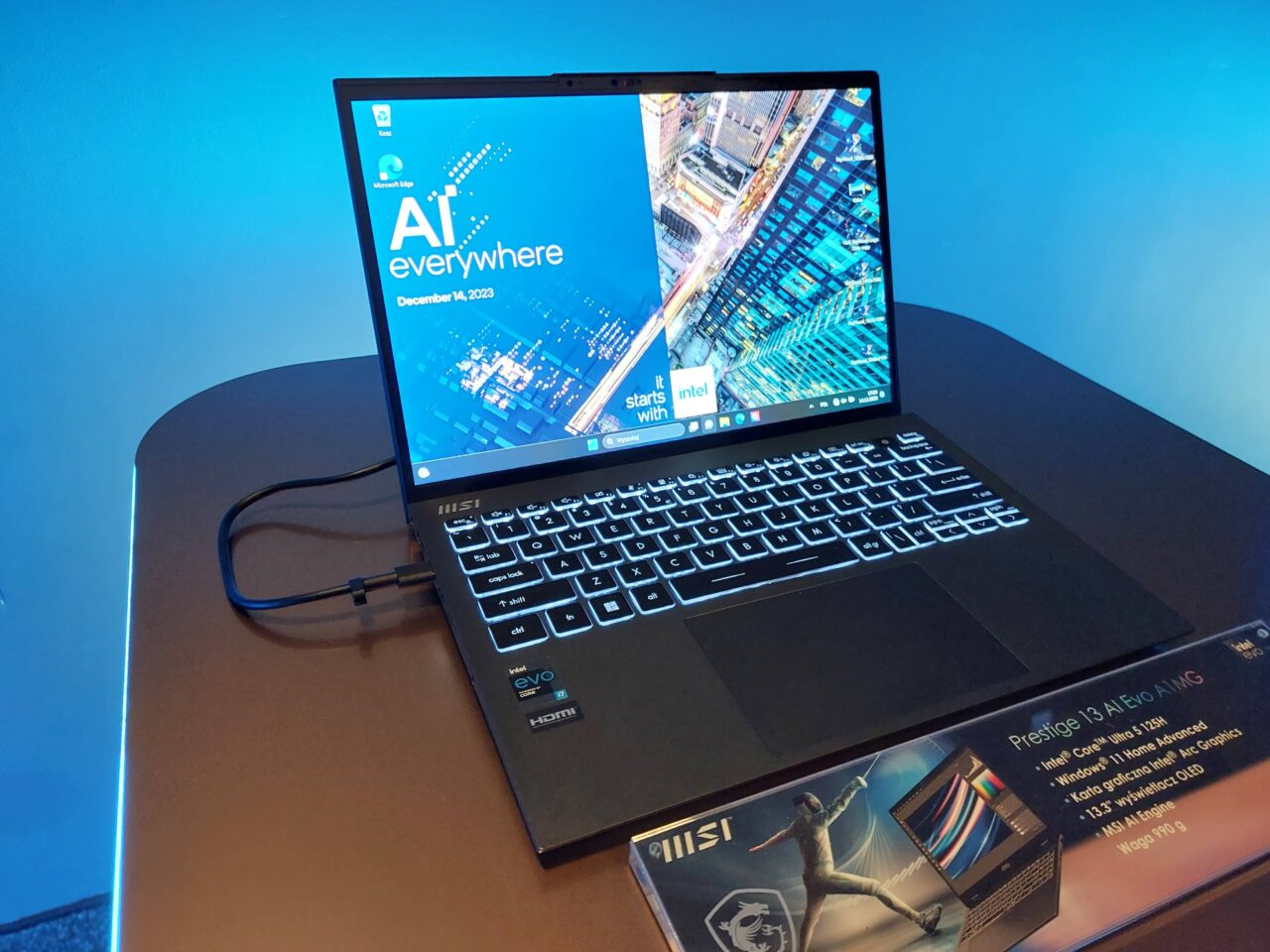 Laptop MSI Prestige 13 AI Evo A1MG z otwartą klapą na stole, wyświetlający tapetę z napisem "AI everywhere" oraz datą 14 grudnia 2023, w tle niebieskie oświetlenie.