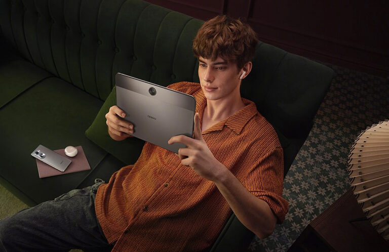 Młody mężczyzna siedzący na zielonej kanapie trzymający tablet OPPO Pad Neo i korzystający z bezprzewodowych słuchawek, obok niego smartfon i ładowarka, w przytulnym, domowym wnętrzu.