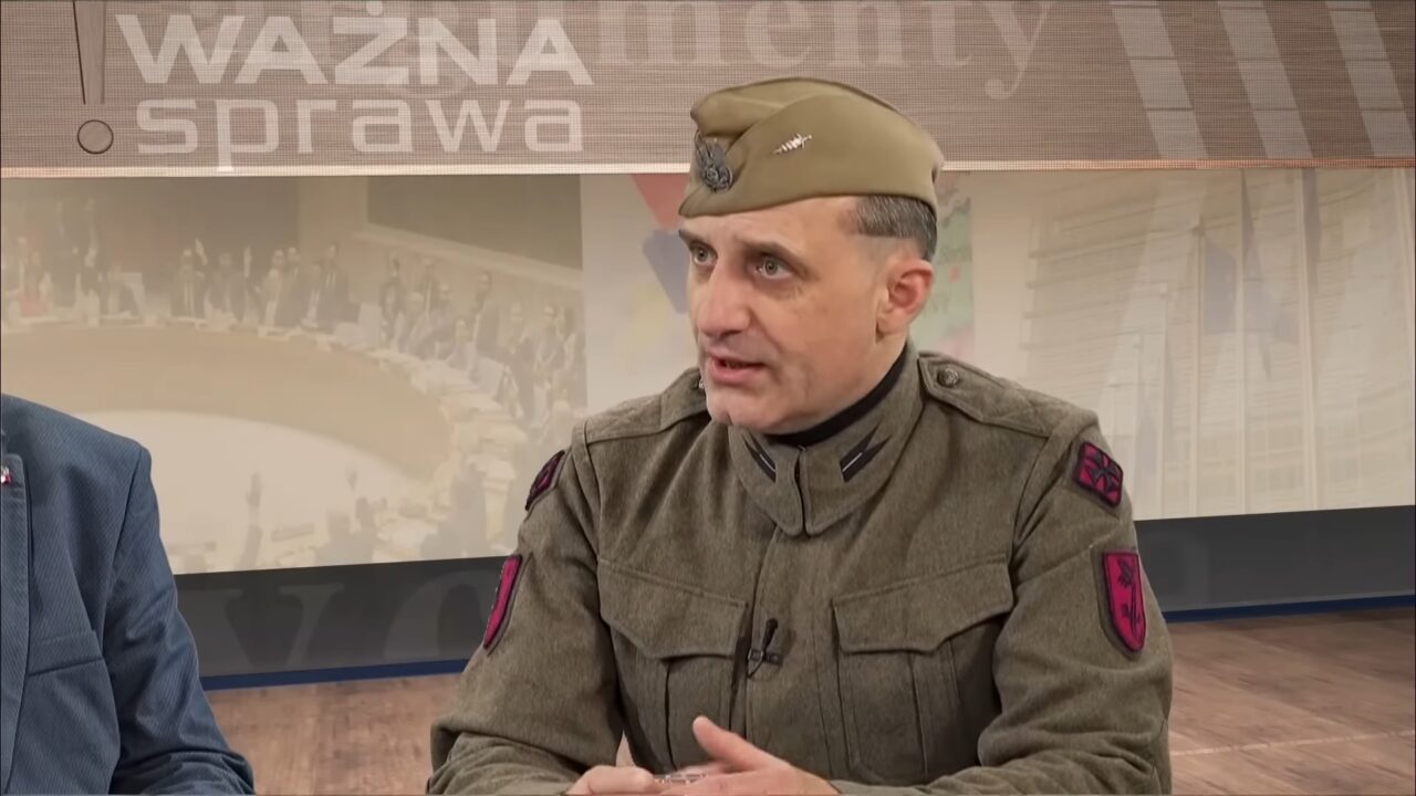 Wojciech Olszański. Mężczyzna ubrany w wojskowy mundur z naszywkami na rękawie, w studiu telewizyjnym z napisem "WAŻNA SPRAWA" w tle.