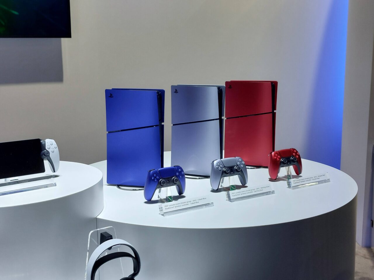 Konsola do gier wideo PlayStation 5 / PS5 w trzech wersjach kolorystycznych: czarnej, niebieskiej i czerwonej, z dopasowanymi kontrolerami, wystawione na białym okrągłym podeście.