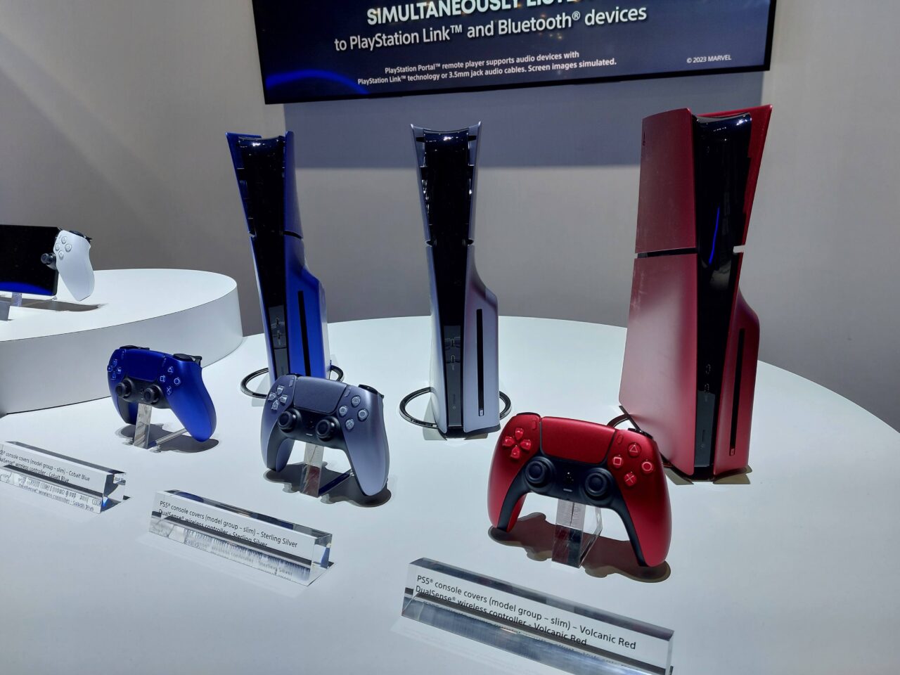 Ekspozycja trzech konsol PlayStation 5 / PS5 i kontrolerów DualSense w kolorach Galatic Purple, Cosmic Red i Midnight Black, umieszczonych na białym stoliku wystawienniczym.