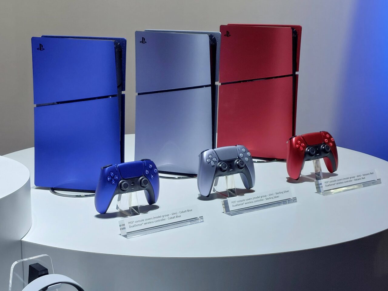Konsola do gier PS5 w różnych kolorach (błękitna, srebrna, czarna, czerwona) i pasujące do nich kontrolery bezprzewodowe, wystawione na białym stoliku wystawienniczym.