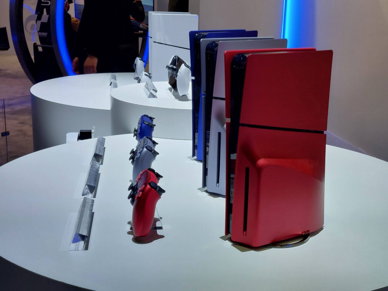 Ekspozycja różnych modeli konsoli do gier PS5 w różnych wersjach kolorystycznych na białym stoliku wystawienniczym.