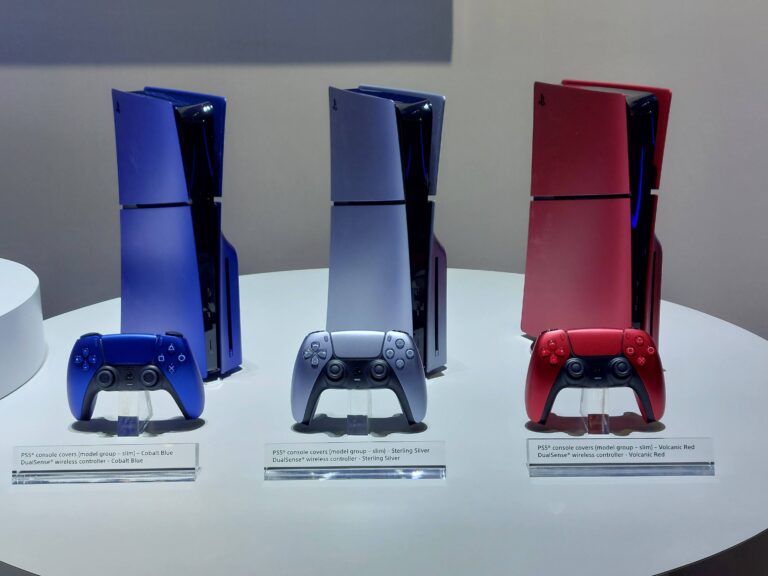 Trzy konsole PlayStation 5 / PS5 w różnych kolorach (kobaltowy niebieski, srebrzysty i wulkaniczny czerwony) z dopasowanymi bezprzewodowymi kontrolerami DualSense prezentowane na wystawie.
