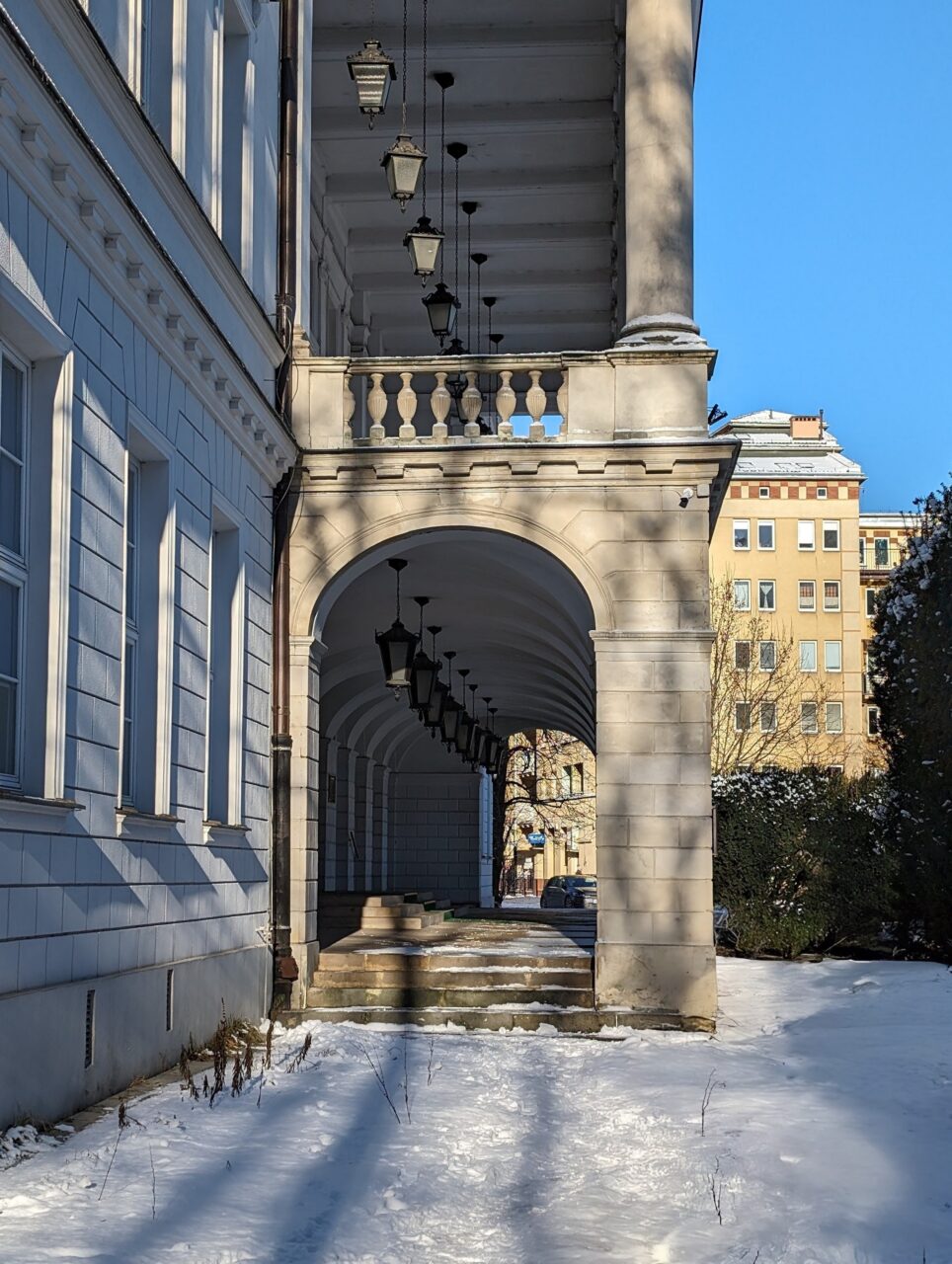 Arkadowy korytarz z klasycznymi latarniami przylegający do białego budynku z widokiem na pokrytą śniegiem ulicę i zabudowania miejskie w tle przy słonecznym dniu.