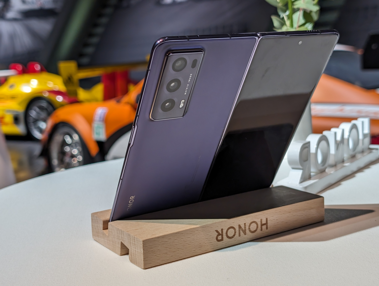 Składany smartfon marki Honor umieszczony na drewnianym stojaku z napisem Honor, z rozmytym tłem przedstawiającym kolorowy samochód wyścigowy.