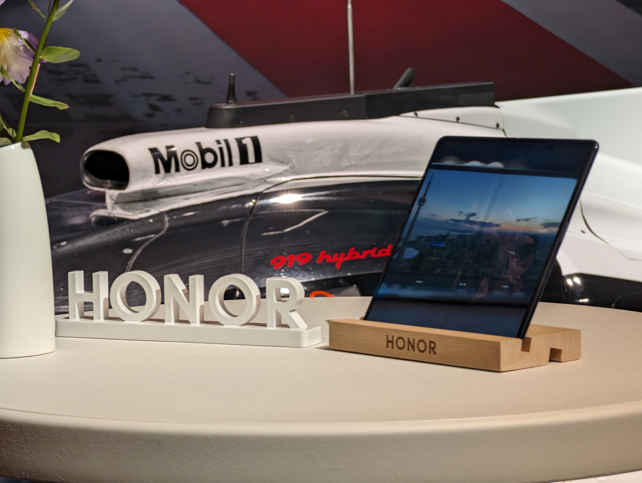 Tablet marki Honor stoi na stojaku z wygrawerowanym logo Honor na pierwszym planie, a w tle część białego pojazdu wyścigowego z napisami Mobil 1 i 919 hybrid.