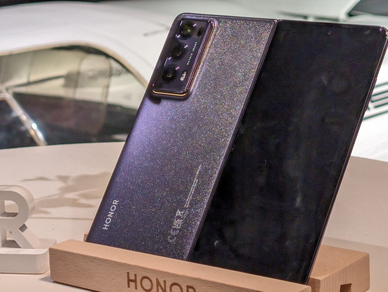 Dwa smartfony marki Honor odbijające światło, z tylnymi aparatami fotograficznymi, wystawione na drewnianym stojaku z logotypem marki.