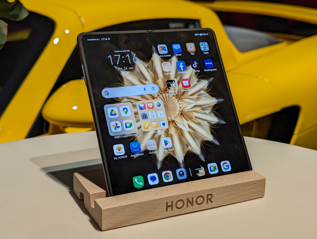 Tablet marki Honor ustawiony na drewnianym stojaku z wyświetlonym ekranem głównym, w tle żółty samochód.