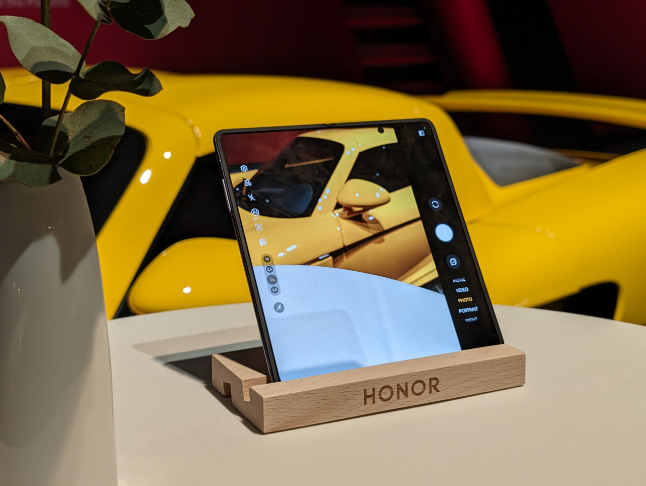 Przenośny tablet marki HONOR stoi na drewnianym stojaku, a na jego ekranie wyświetla się obraz żółtego samochodu sportowego; w tle widoczna część doniczki z rośliną.