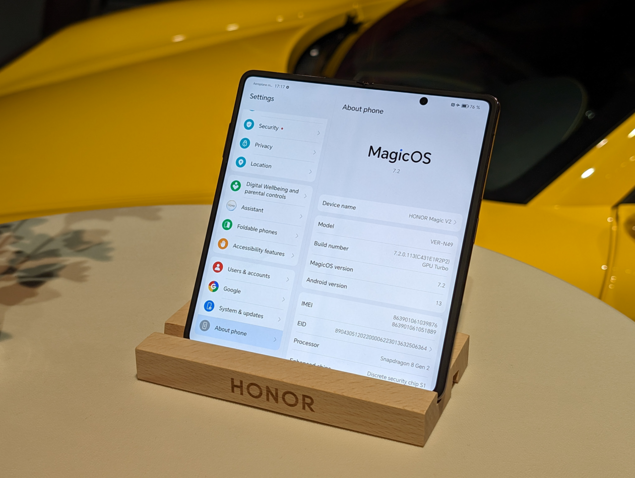 Smartfon Honor umieszczony na drewnianym podstawku z otwartymi ustawieniami systemowymi na ekranie, na tle żółtego obiektu.