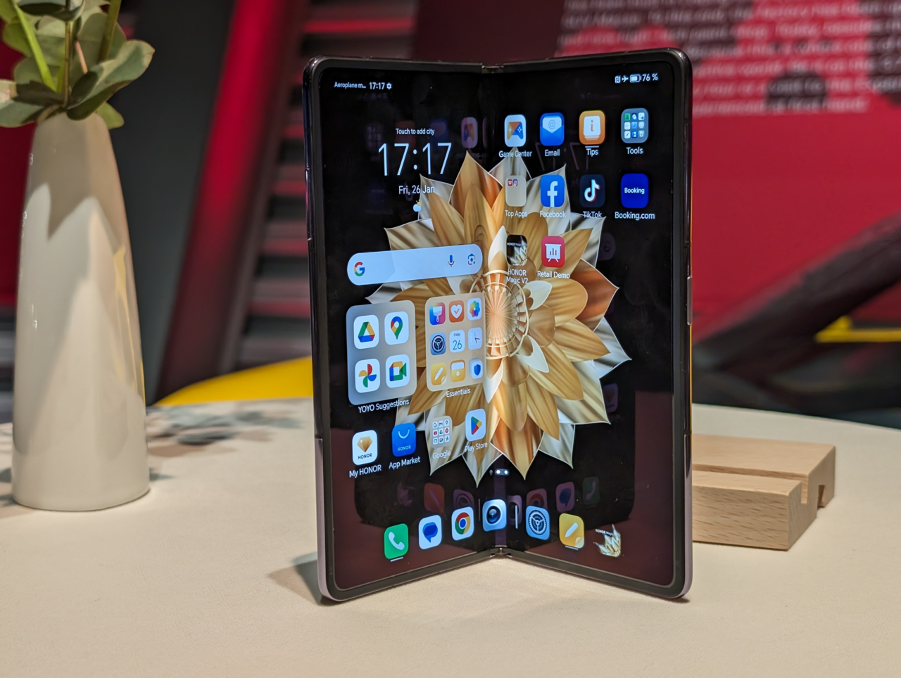 Składany smartfon stoi na stole obok doniczki z rośliną, z wyświetlonym ekranem głównym pełnym ikon aplikacji.