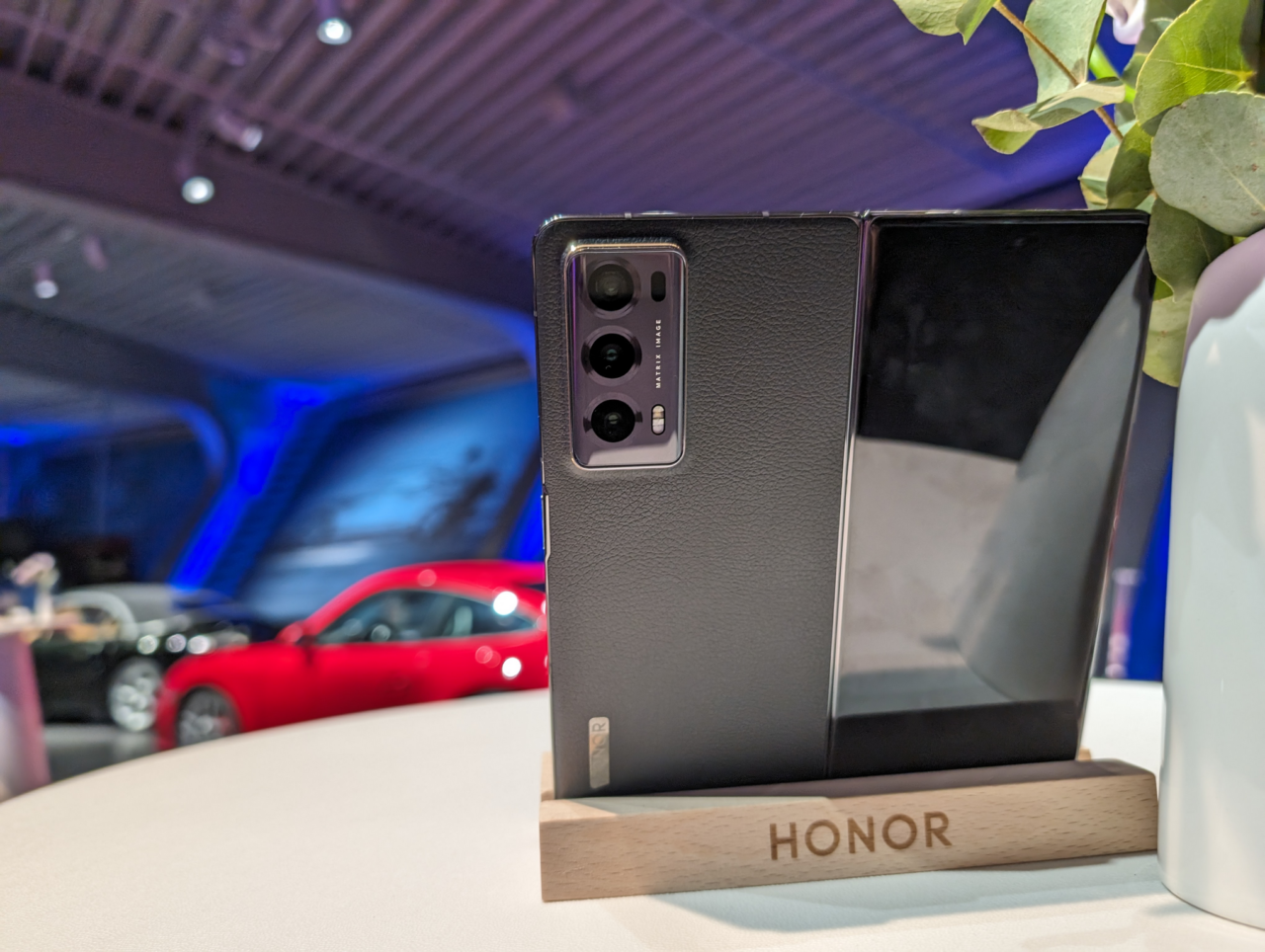 Smartfon marki Honor umieszczony na podstawce, przedzielony na półekranowy wyświetlacz z widocznym odbiciem, na tle rośliny i rozmazanego czerwonego samochodu.