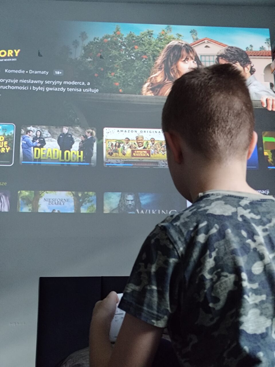 Chłopiec w moro ogląda telewizję, na ekranie interfejs platformy streamingowej z filmami i serialami.