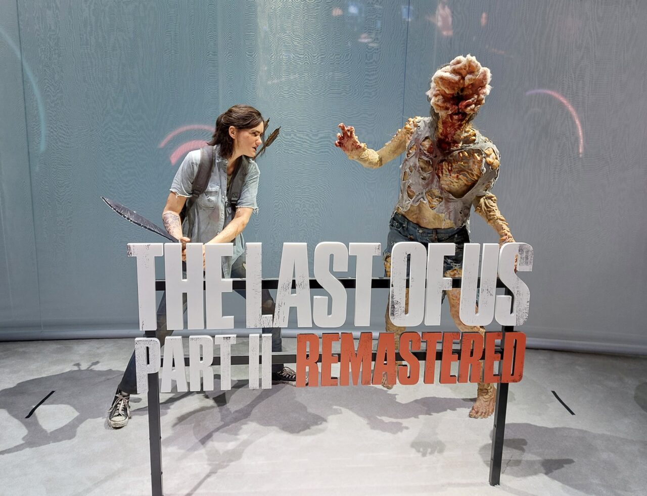 Postać ludzka trzymająca nóż przeciwko stworzeniu przypominającemu zombie, na tle napisu "THE LAST OF US PART II REMASTERED". Promocja gry na PS5