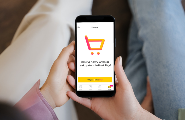 Osoba trzymająca smartfon z ekranem prezentującym aplikację InPost Pay do zakupów z napisem "Odkryj nowy wymiar zakupów z InPost Pay!" i ikoną żółtego wózka na górze.