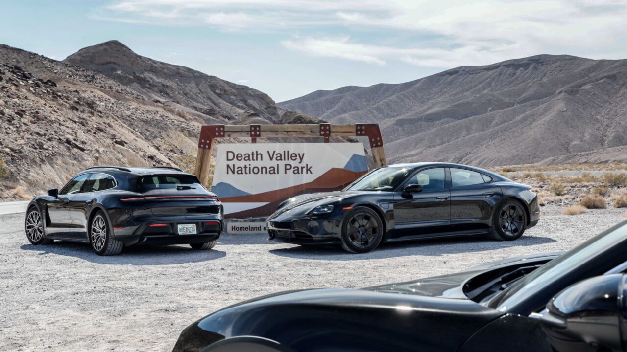 Porsche Taycan. Dwa czarne sportowe samochody zaparkowane obok siebie przed brązowym znakiem "Death Valley National Park" na tle skalistego pustynnego krajobrazu.