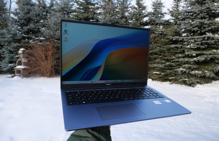 Laptop marki Huawei trzymany na tle zimowego ogrodu z drzewami iglastymi i śniegiem na ziemi.