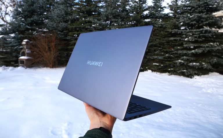 Otwarty laptop HUAWEI MateBook D 16 trzymany w ręku. W tle rozmazany śnieg i drzewa iglaste.