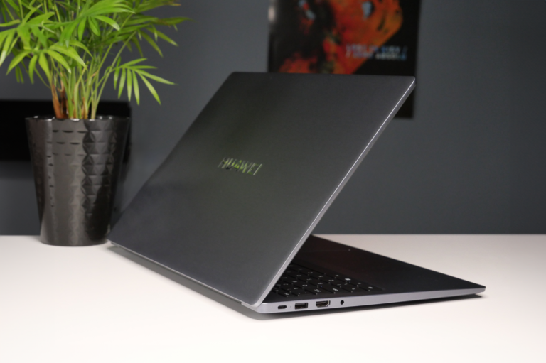 Grafitowy laptop marki Huawei na biurku, częściowo zamknięty, z logo producenta na pokrywie, w tle czarny doniczka z zieloną rośliną i plakat na ścianie.