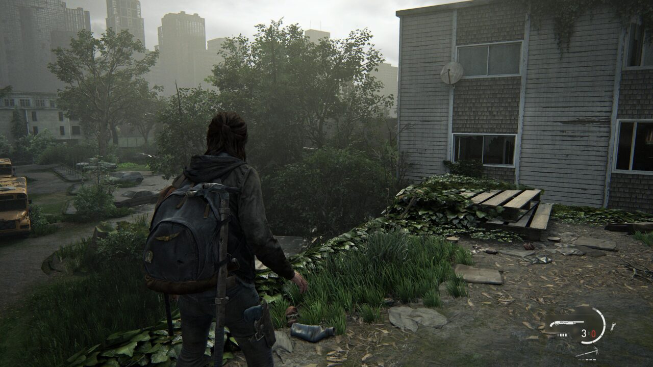 Postać kobieca z plecakiem stoi na zewnątrz opuszczonego budynku, patrząc na zarośnięte ulice z porozrzucanymi samochodami i autobusem szkolnym w szary, pochmurny dzień. Screen z gry The Last of Us Part II Remastered na PS5.