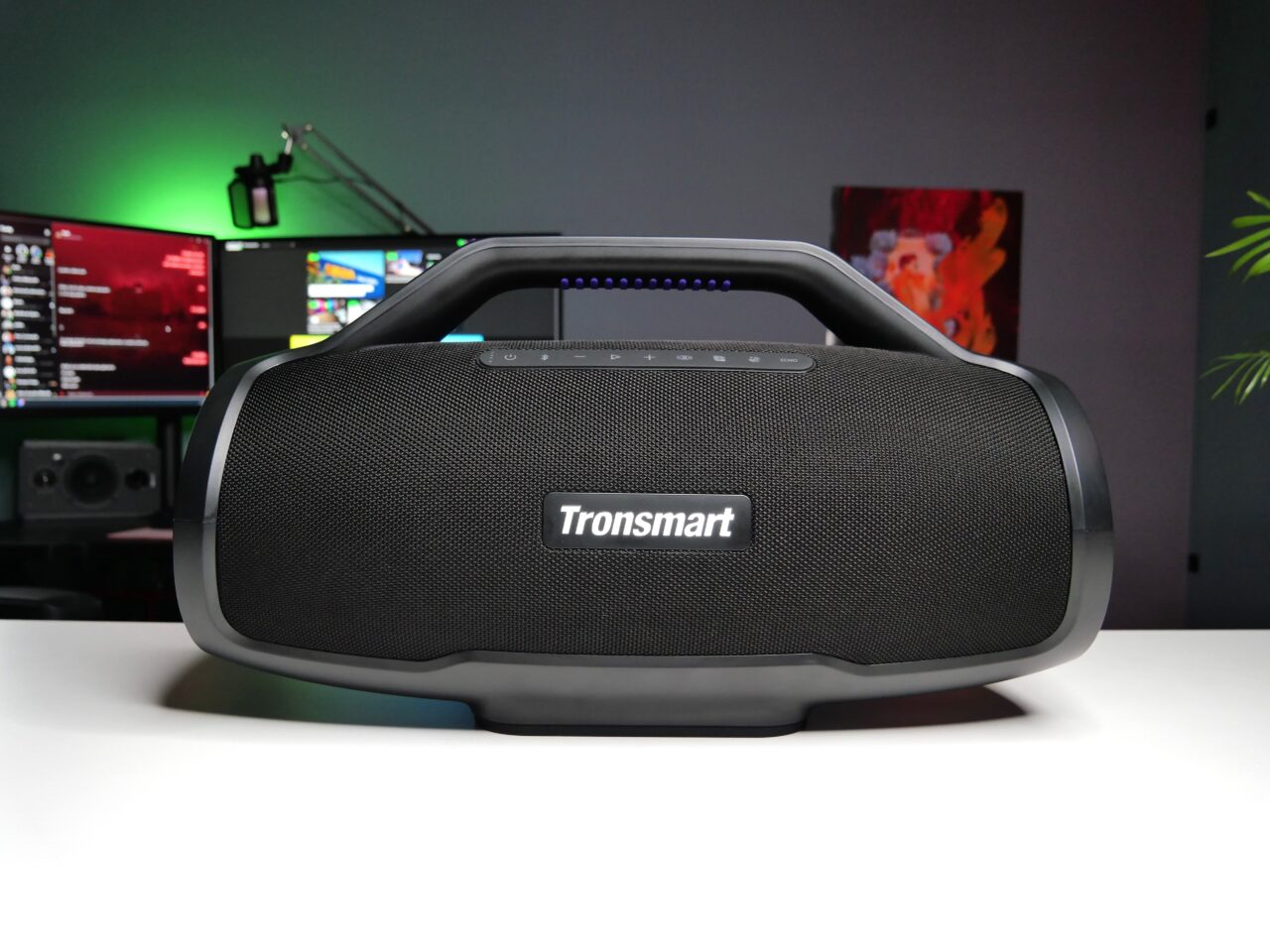 Czarny, przenośny głośnik Bluetooth marki Tronsmart na biurku, w tle komputer z włączonymi monitorami i kolorowe oświetlenie RGB.