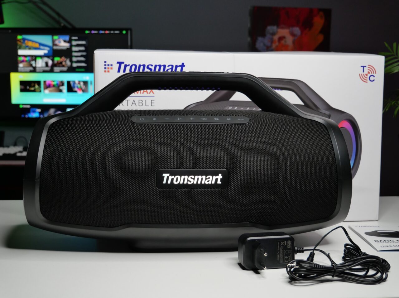 Przenośny głośnik Bluetooth marki Tronsmart na biurku, z tyłu kartonowe opakowanie, na pierwszym planie zasilacz i przewód.