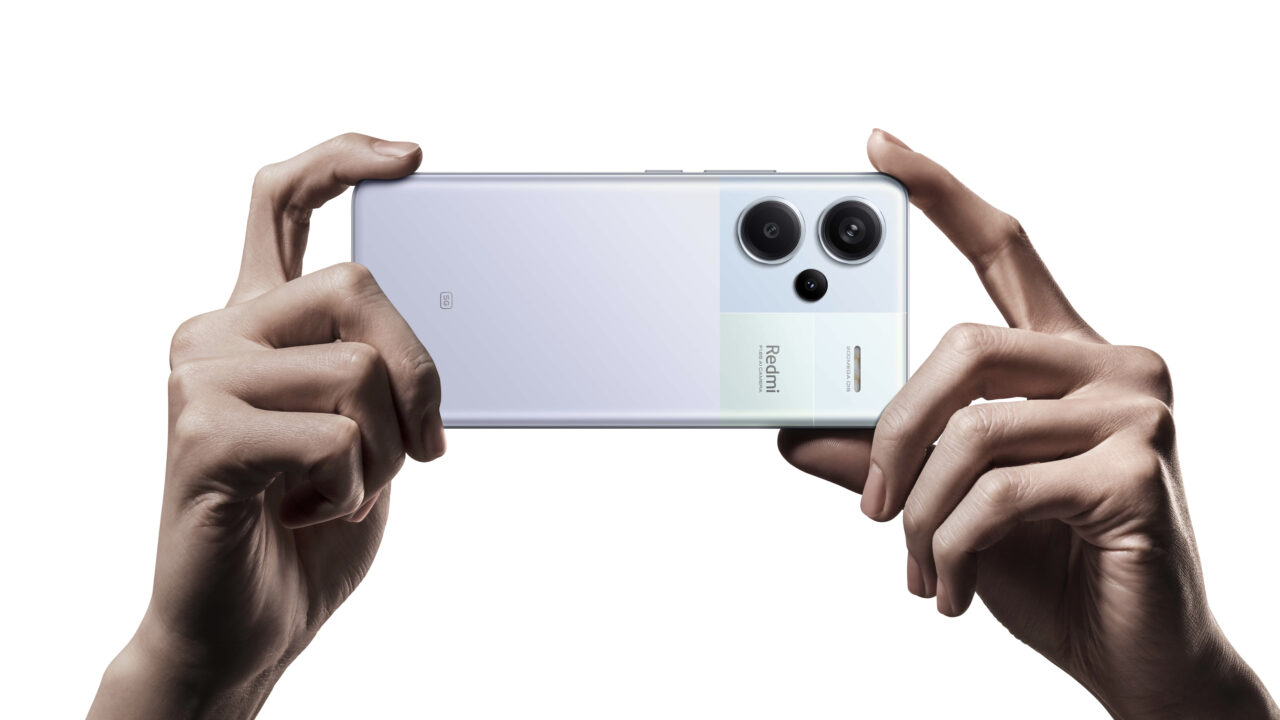 Zdjęcie smartfona Redmi Note 13 Pro+ 5G trzymanego poziomo w dwóch dłoniach z widocznym podwójnym aparatem fotograficznym i logo marki.
