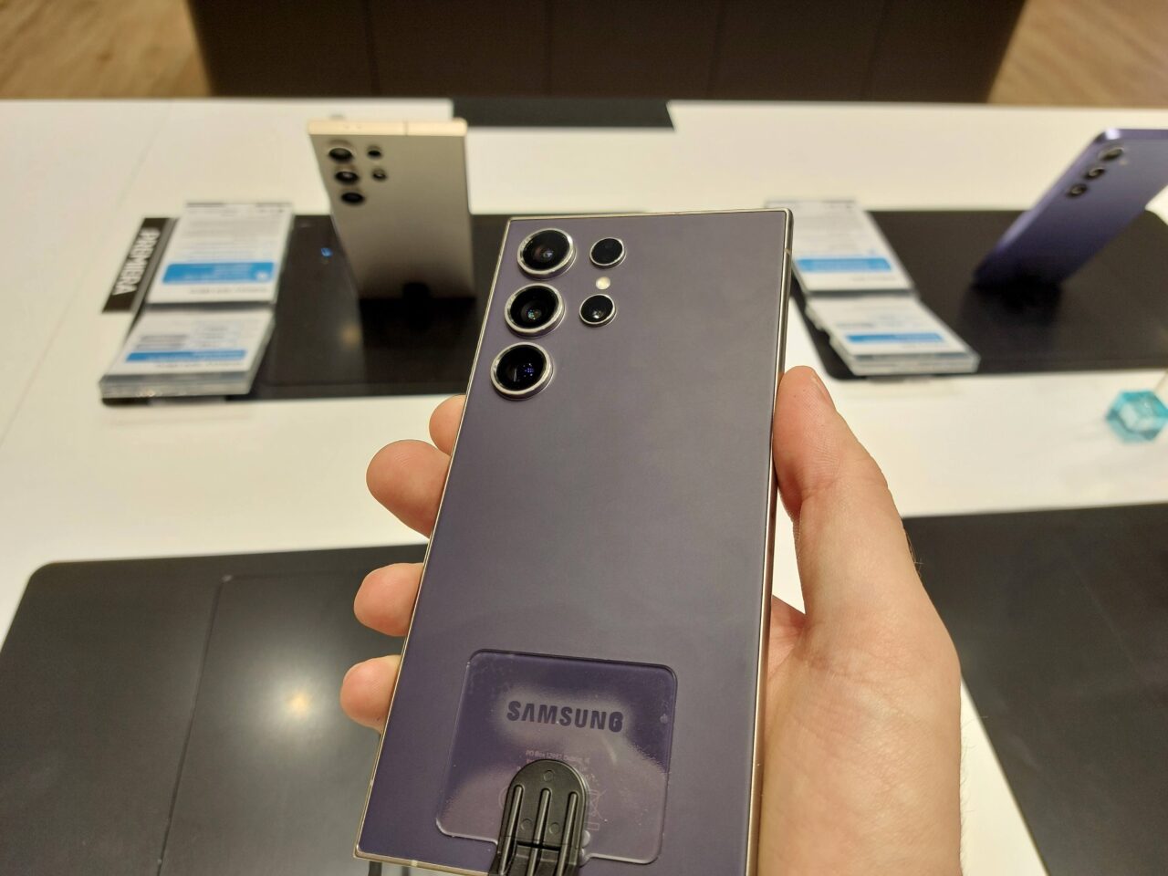 Osoba trzyma tylną część smartfona Samsung z pięcioma aparatami w sklepie elektronicznym, z wystawionymi telefonami w tle.