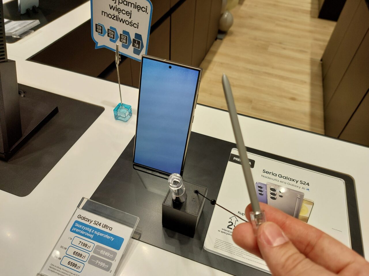 Smartfon Galaxy S24 Ultra wystawiony w sklepie, zabezpieczony przeciwdziałającym kradzieży urządzeniem, obok karty informacyjnej z ceną i specyfikacją oraz długopisem dotykowym trzymanym przez osobę robiącą zdjęcie.