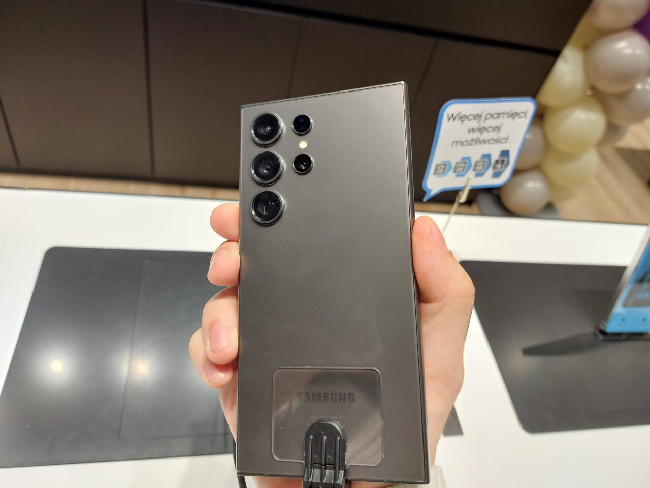 Czarna tylna część smartfona Samsung z pięcioma obiektywami kamery trzymana w dłoni, w tle fragment sklepu z reklamą o większej pamięci i wydajności.