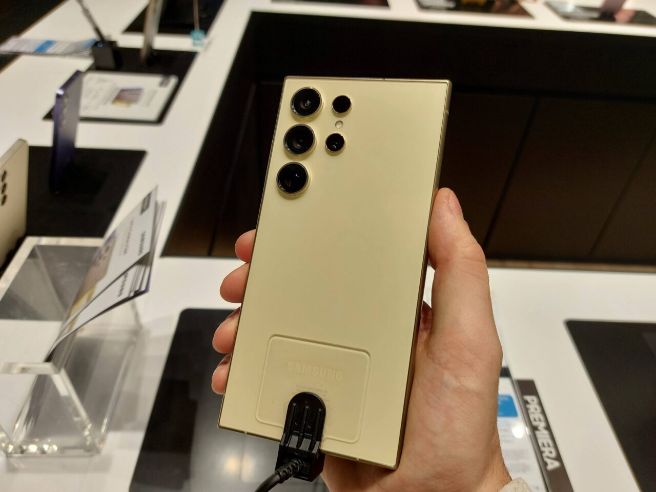 Ręka trzymająca złoty smartfon Samsung z pięcioma obiektywami aparatu ustawionymi w dwóch rzędach, podłączonego kablem do ekspozycji w sklepie.