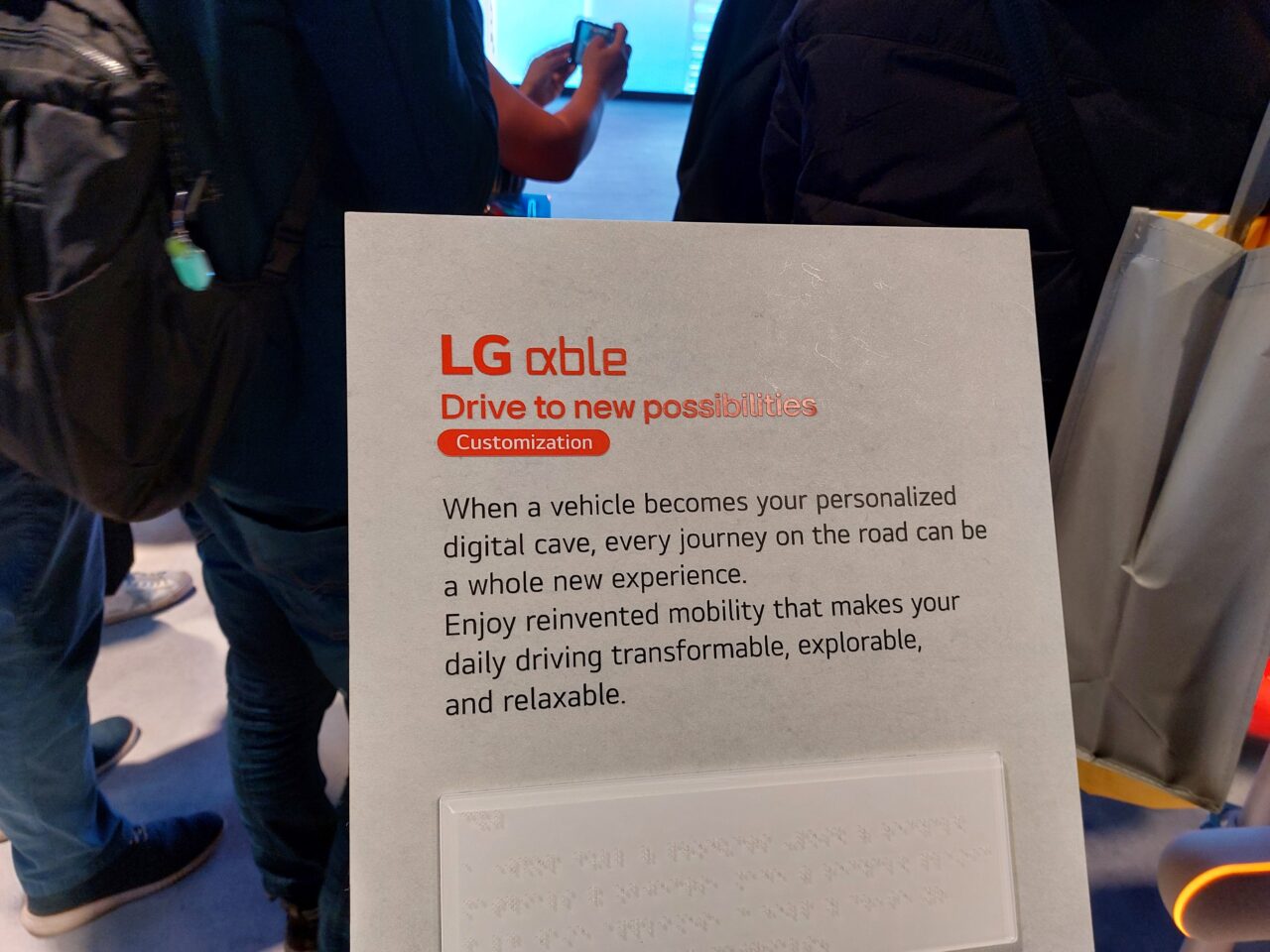 Plakat reklamowy firmy LG z sloganem "LG xable Drive to new possibilities Customization" i opisem produktu, z tłumem osób w tle.