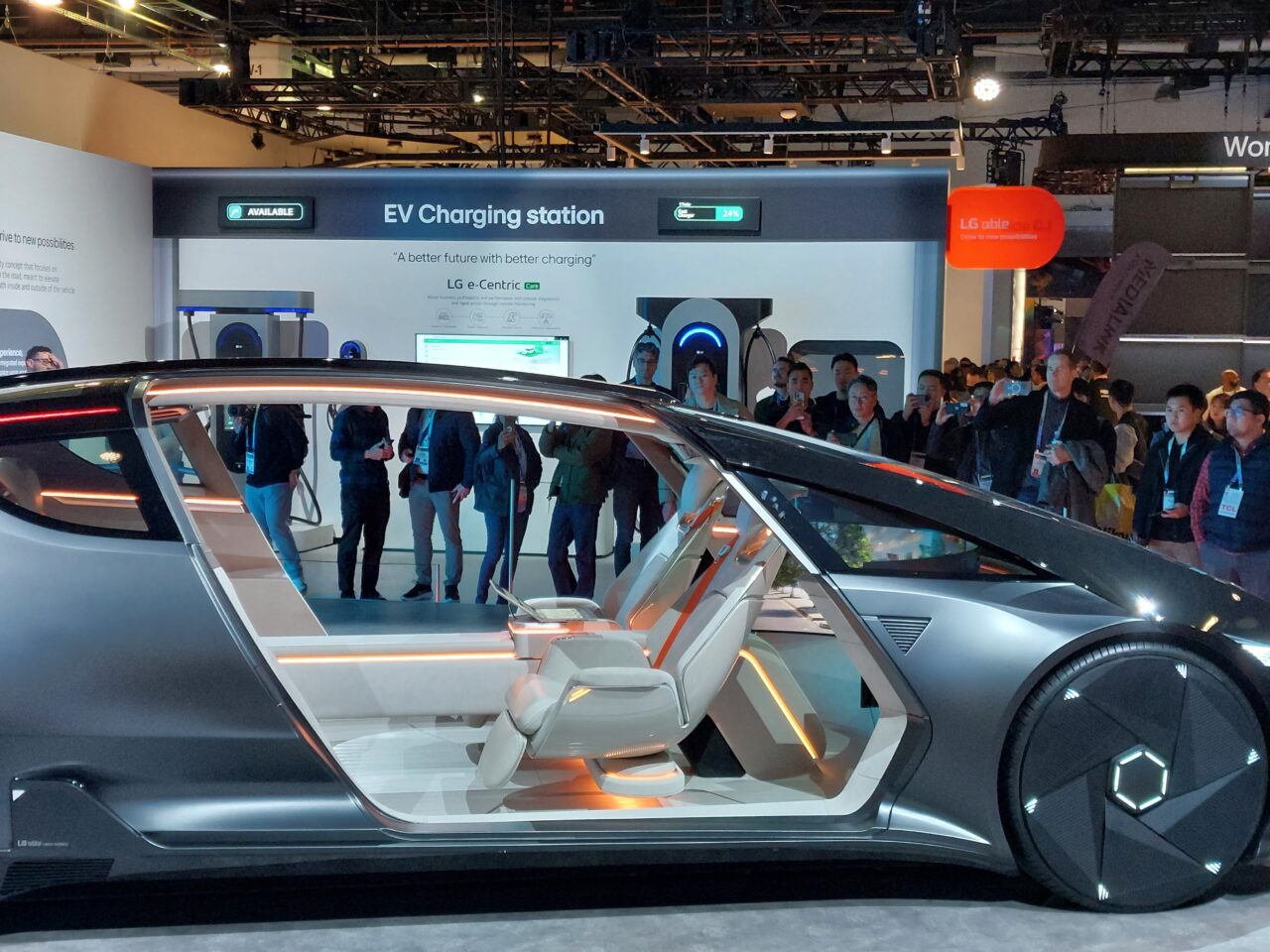 Ekspozycja nowoczesnego elektrycznego samochodu koncepcyjnego z otwartymi drzwiami oraz grupa ludzi oglądających pojazd i stację ładowania EV w tle.