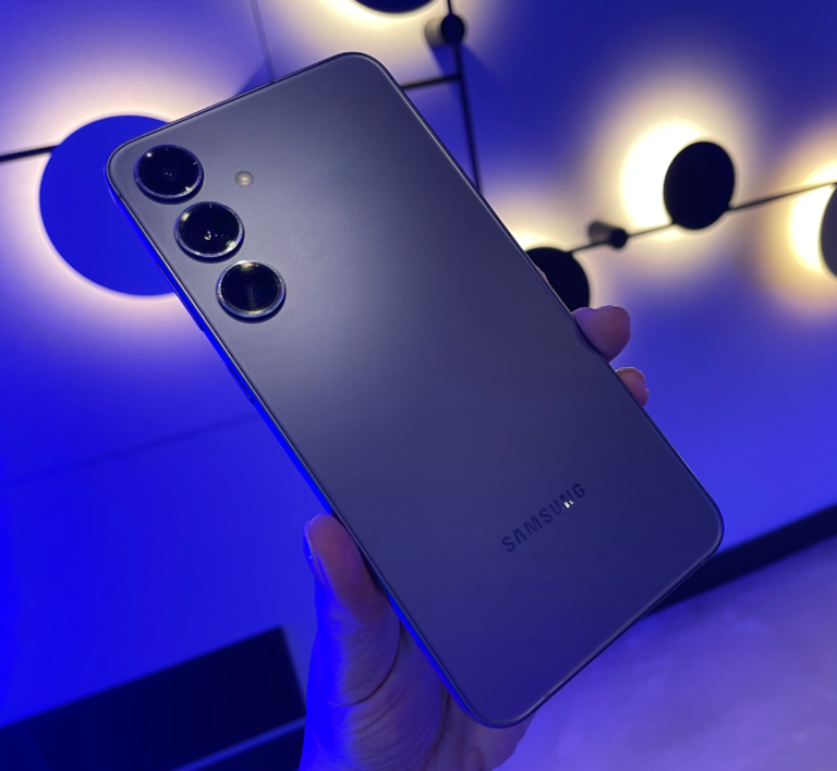 Smartfon marki Samsung trzymany w dłoni na tle niebieskiego oświetlenia, z widocznym tylnym aparatem z trzema obiektywami.