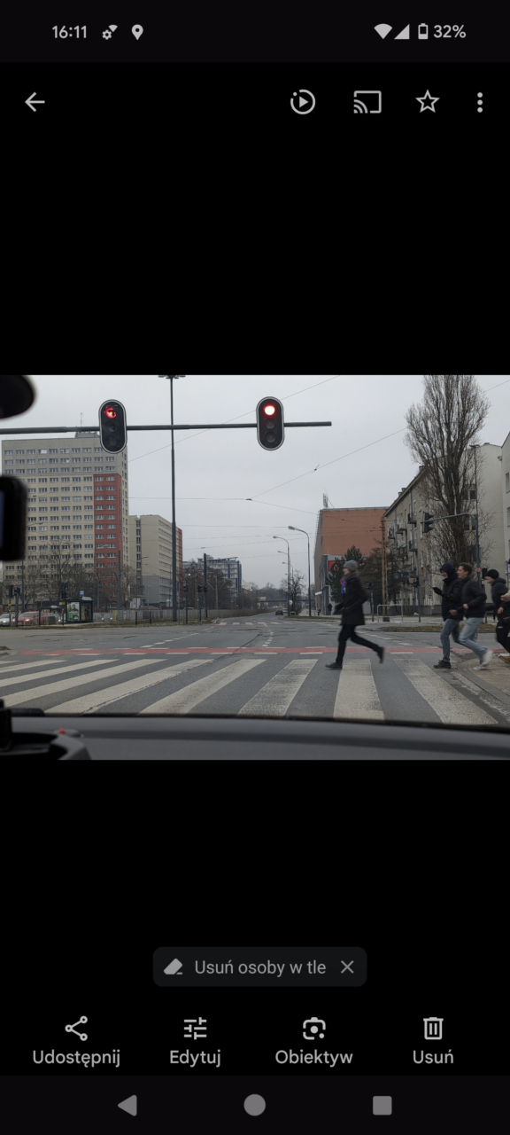 Zdjęcie wykonane z wnętrza samochodu przedstawiające miejskie skrzyżowanie z sygnalizacją świetlną wskazującą czerwone światło oraz pieszych przechodzących przez przejście dla pieszych. W tle widoczne są miejskie budynki i gołe drzewa. Na pierwszym planie fragment deski rozdzielczej samochodu.