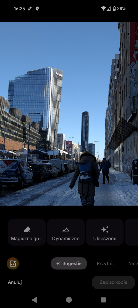 Zdjęcie ulicy w mieście z wysokimi nowoczesnymi wieżowcami i chodnikiem pokrytym śniegiem, po którym idą ludzie, widok w słoneczny dzień.