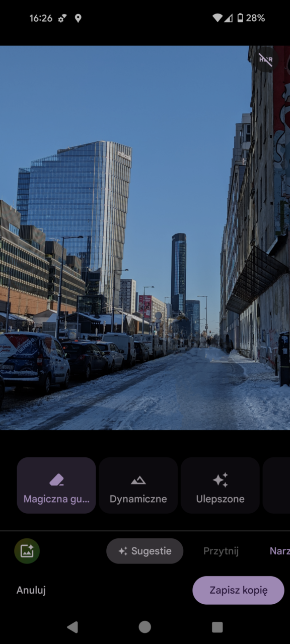 Ulica w miejskiej dzielnicy z wysokimi, nowoczesnymi budynkami i drogą pokrytą śniegiem, prezentowana na ekranie smartfona z widocznymi opcjami edycji zdjęć.