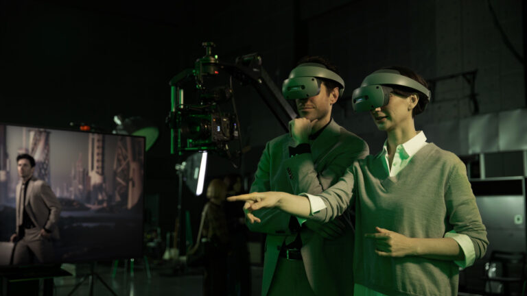 Dwóch ludzi w zielonych ubraniach używa gogli do rzeczywistości wirtualnej w studiu filmowym, w tle widoczny jest wyświetlacz i profesjonalny sprzęt filmowy.